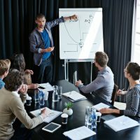 Shot of a young man giving a presentation on a whiteboard to colleagues sitting around a table in a boardroom.