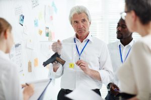 Mature forensic examiner showing gun taken from crime scene to his colleagues while standing at investigation board and participating in discussion